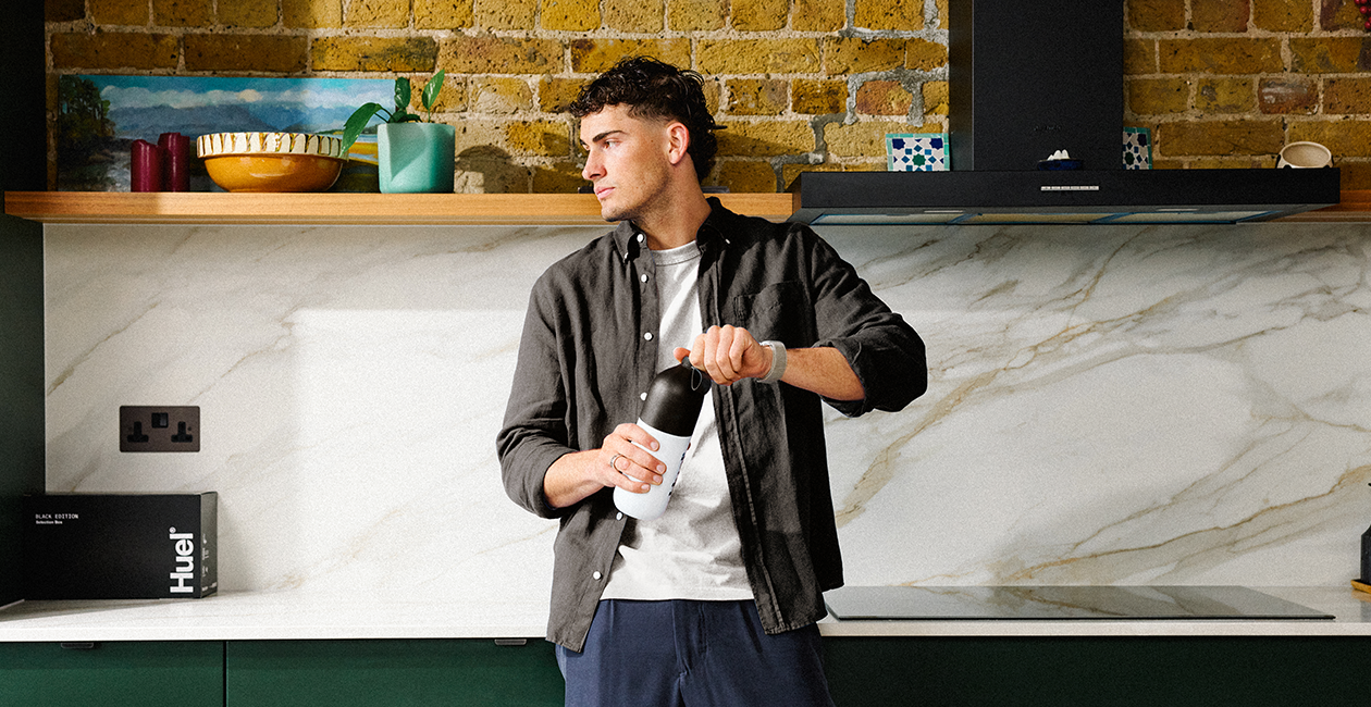 A man holding a Huel shaker with Huel in the background, leaning on a kitchen worktop
