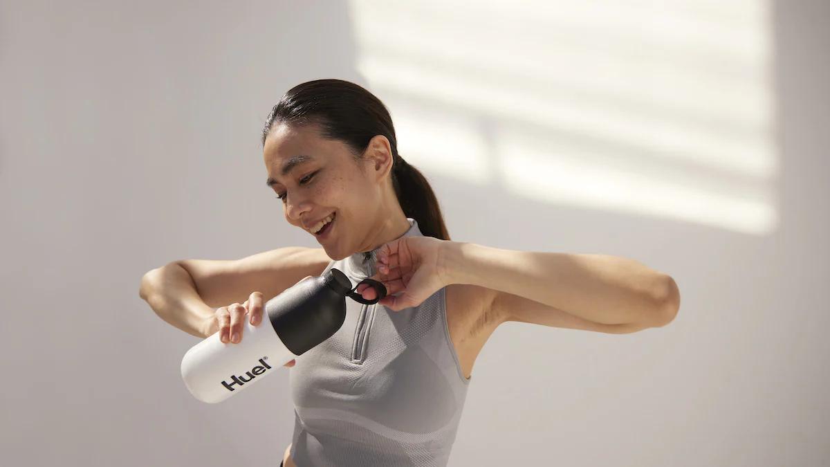 woman wearing fitness gear and smiling while opening the lid on her huel shaker