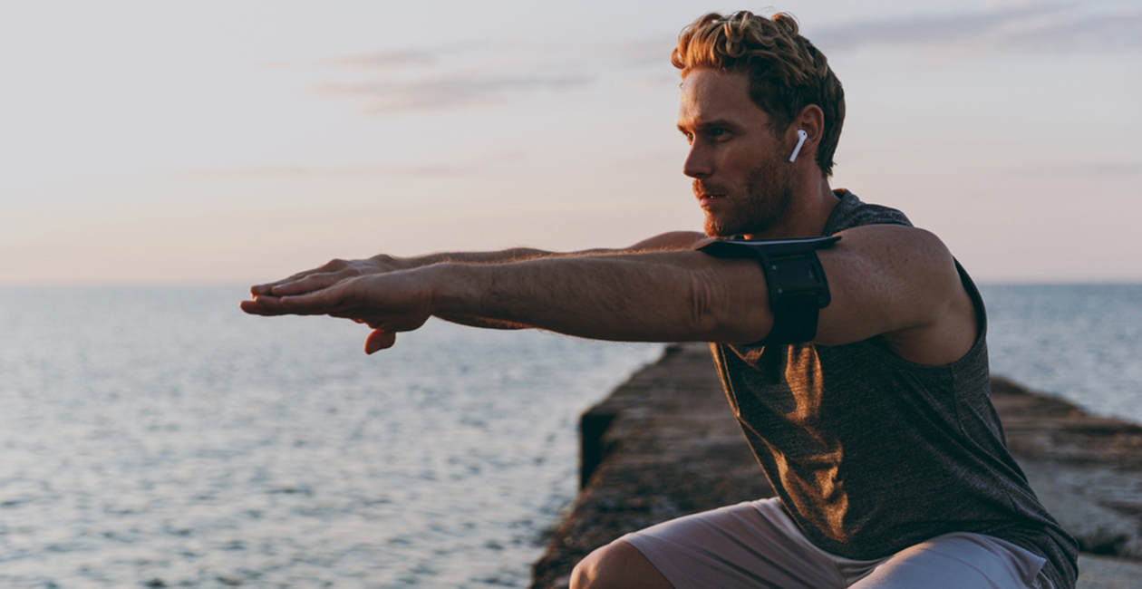 A man in activewear and earphones in mid squat with a sunset in the background