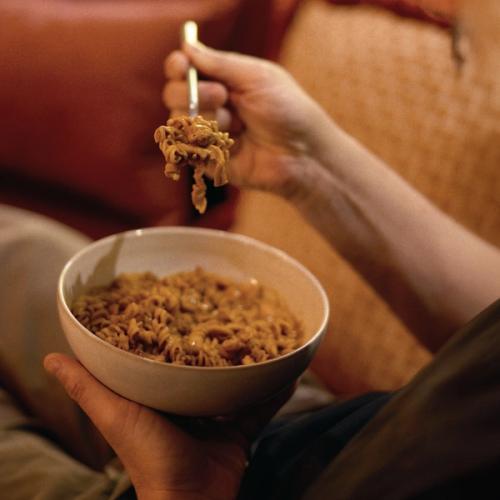 A close up lifestyle photo of a person holding a bowl of pasta while lifting a forkful. The warm lighting suggests a comforting ready to eat meal moment at home.