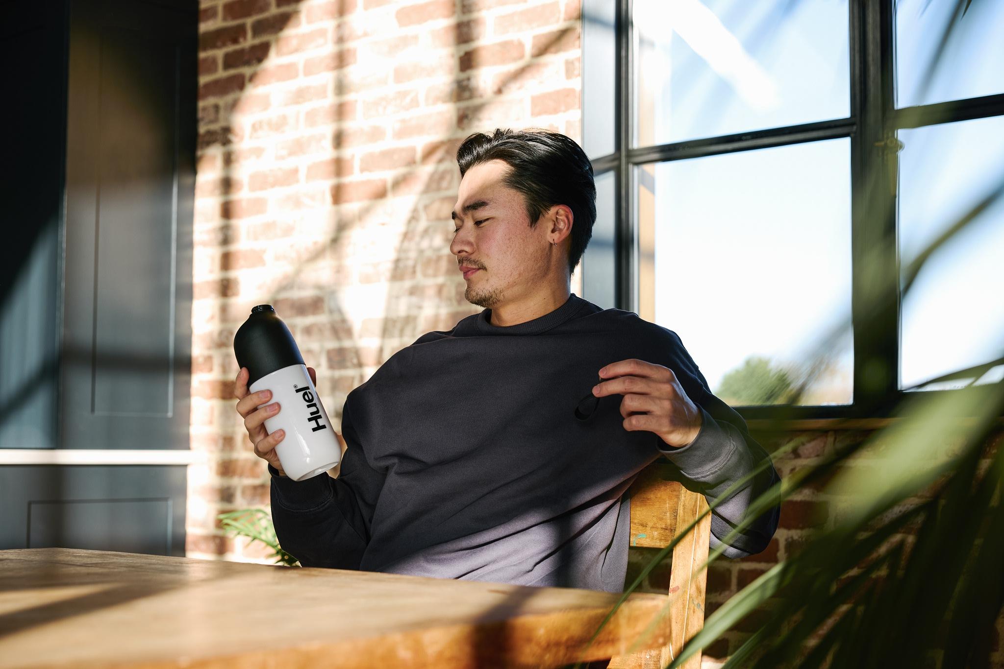 man sat on a chair at a table by a window holding a huel shaker with the sun shining through