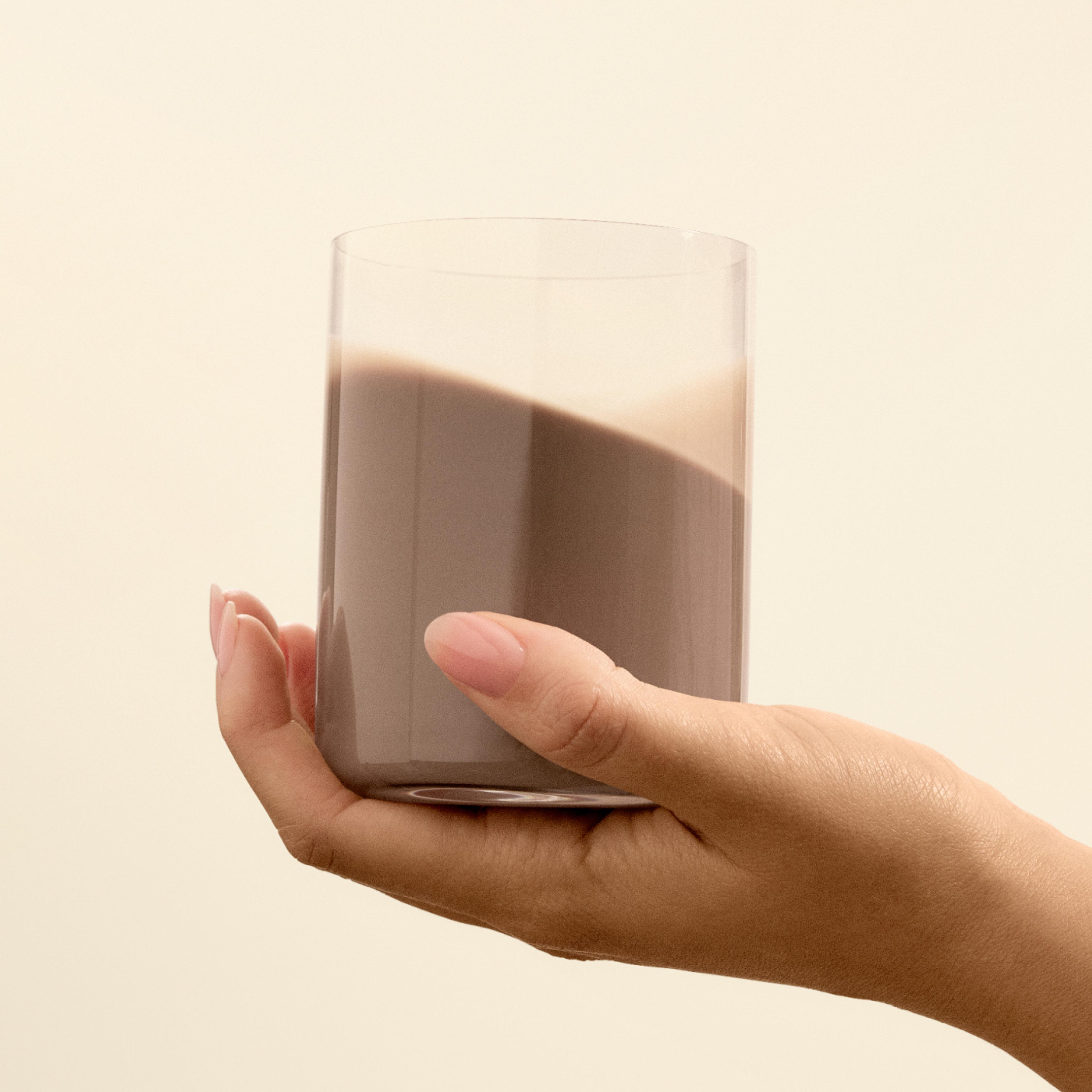 A glass being held by a hand with chocolate flavoured Huel ready-to-drink lite in it on a beige background