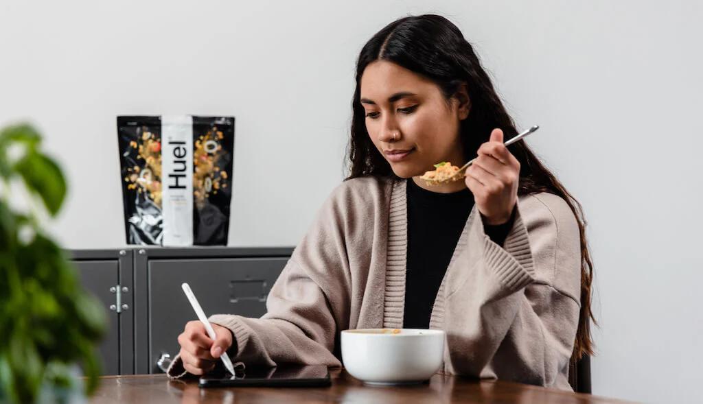 Woman eating hot & savoury from a bowl with a Huel pouch in the background