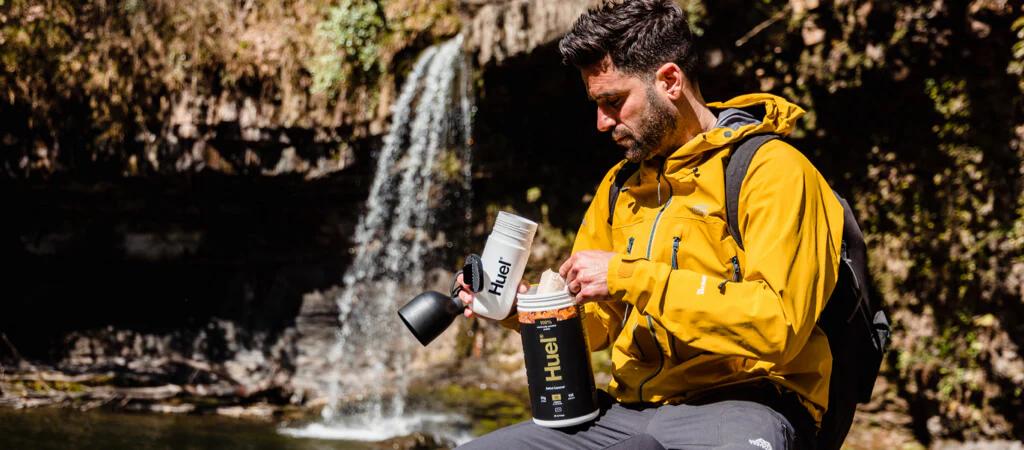 Man wearing a yellow jacker using Huel Complete Protein whilst sat with a waterfall in the background