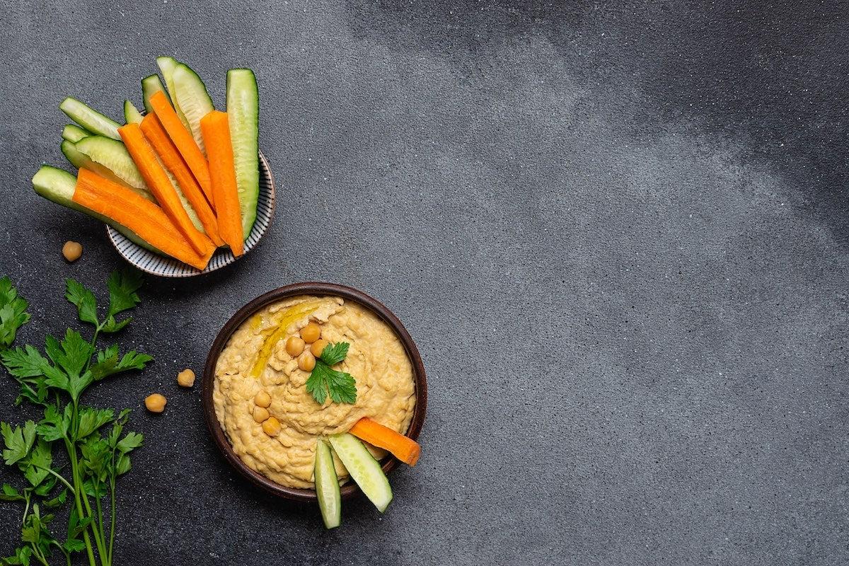 Carrot and cucumber sticks in a bowl and hummus with some chickpeas on top in another bowl
