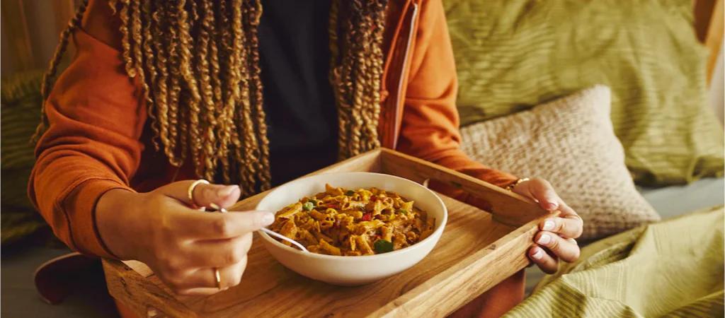 Image of a woman eating Huel Hot & Savoury pasta from a bowl on a rustic tray on a green sofa