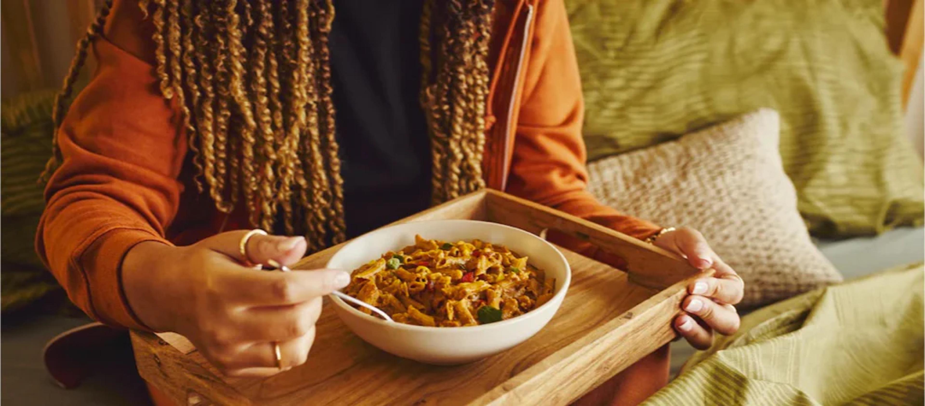 Image of a woman eating Huel Hot & Savoury pasta from a bowl on a rustic tray on a green sofa