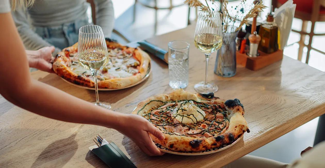 two traditional stone baked pizzas being served to a table at a restaurant with two glasses of white wine on the table