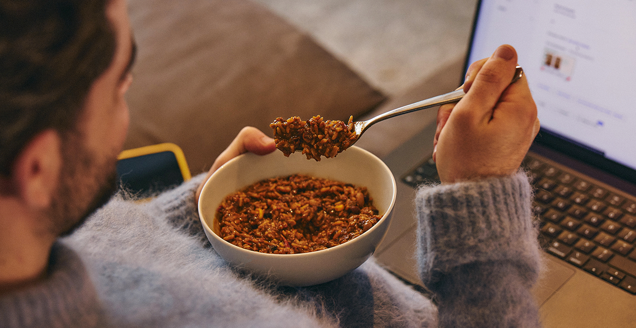 person eating Huel hot & savoury from a bowl looking at their laptop