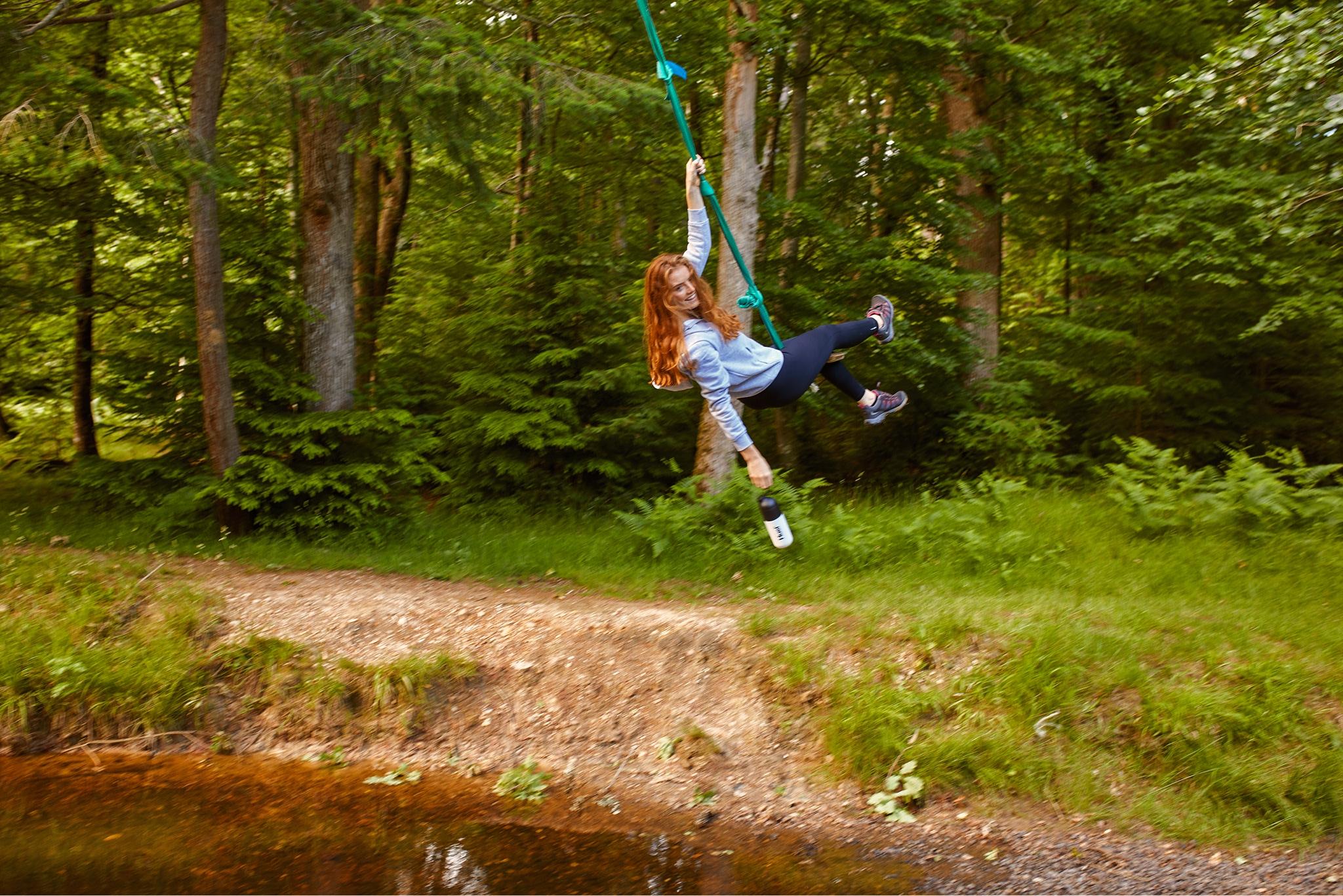 woman swinging on a rope from a tree outside holding a huel shaker