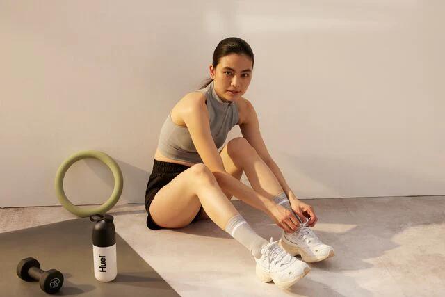 woman sat on the floor tying her shoelace with a dumbbell and huel shaker on a yoga mat next to her