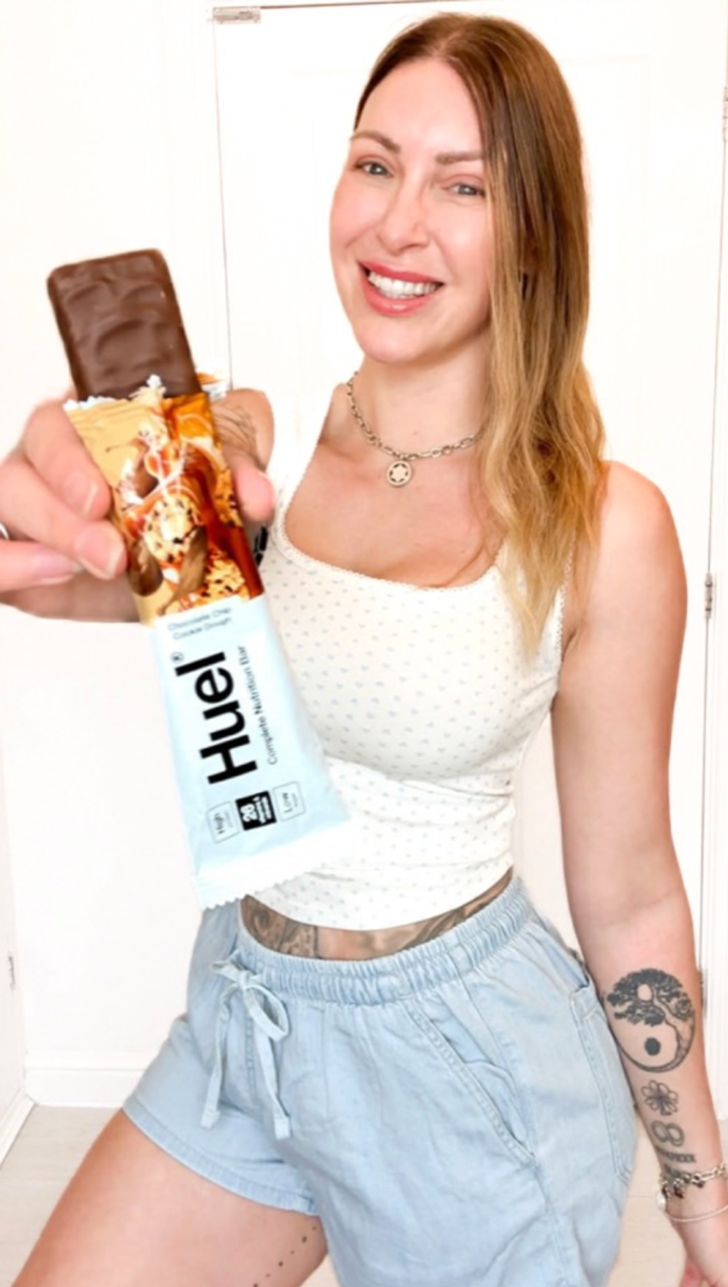 A lady wearing a black t-shirt and shorts smiling holding a Huel shaker