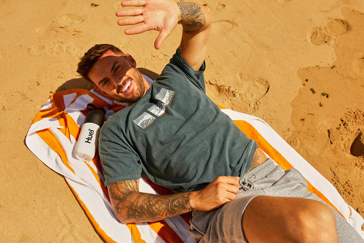 Man laying on a towel on a beach in the sun with a Huel shaker next to him
