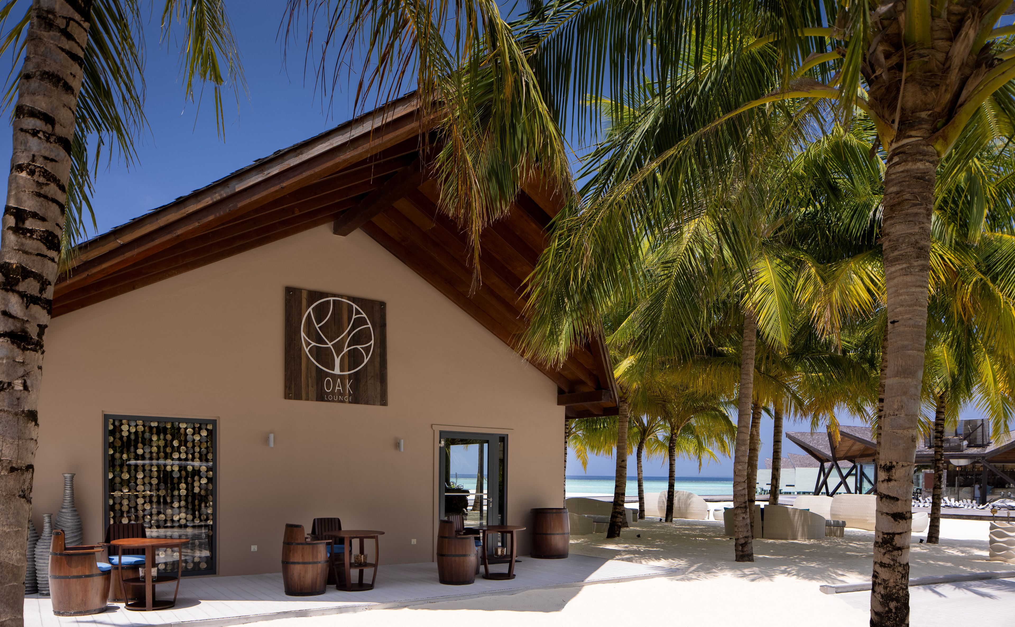 a building with barrels and chairs in front of it is surrounded by palm trees on a beach .