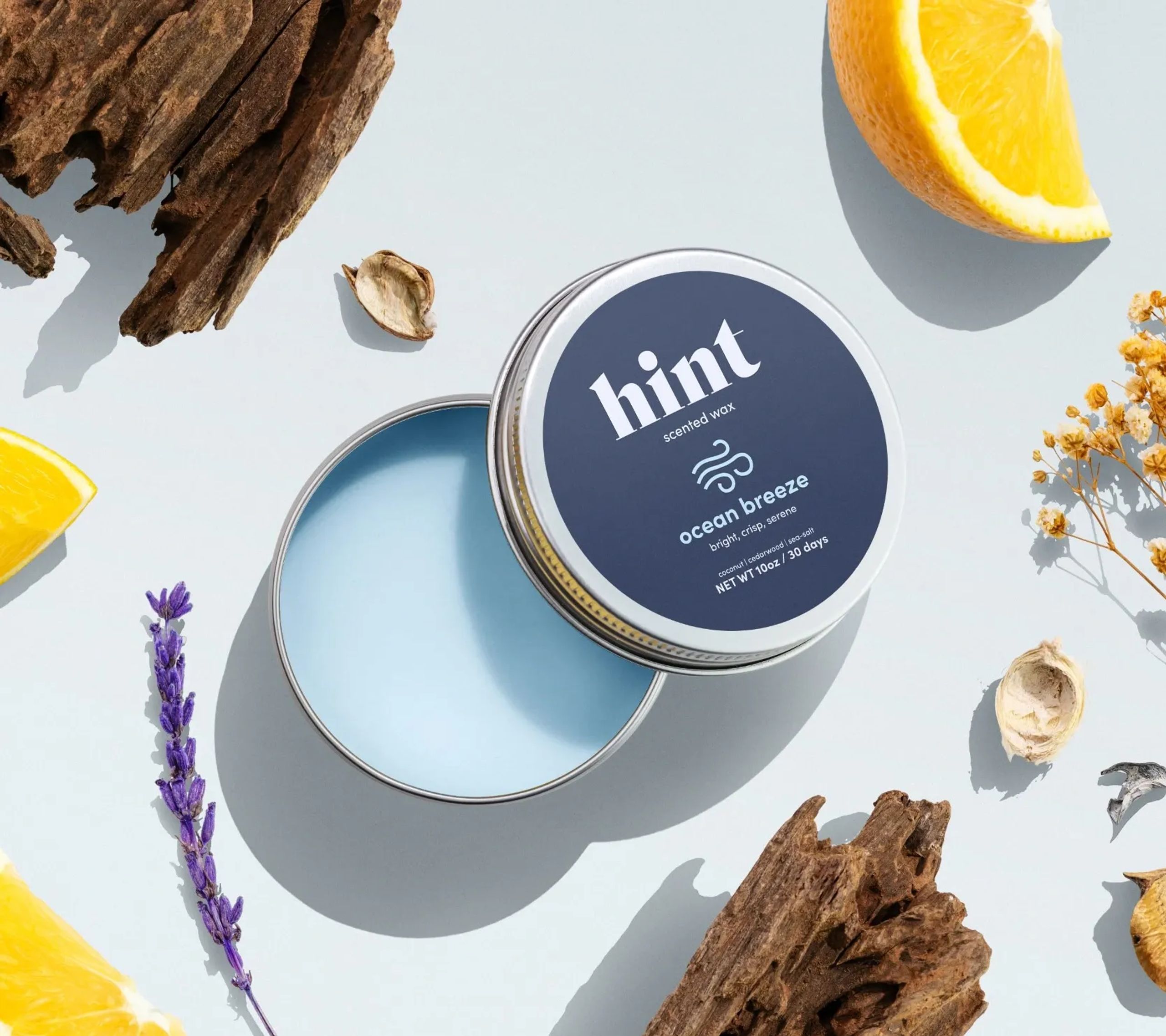 Flat-lay photo of Hint’s “Ocean Breeze” scented wax tin, surrounded by natural elements like orange slices, lavender, and driftwood on a light background.