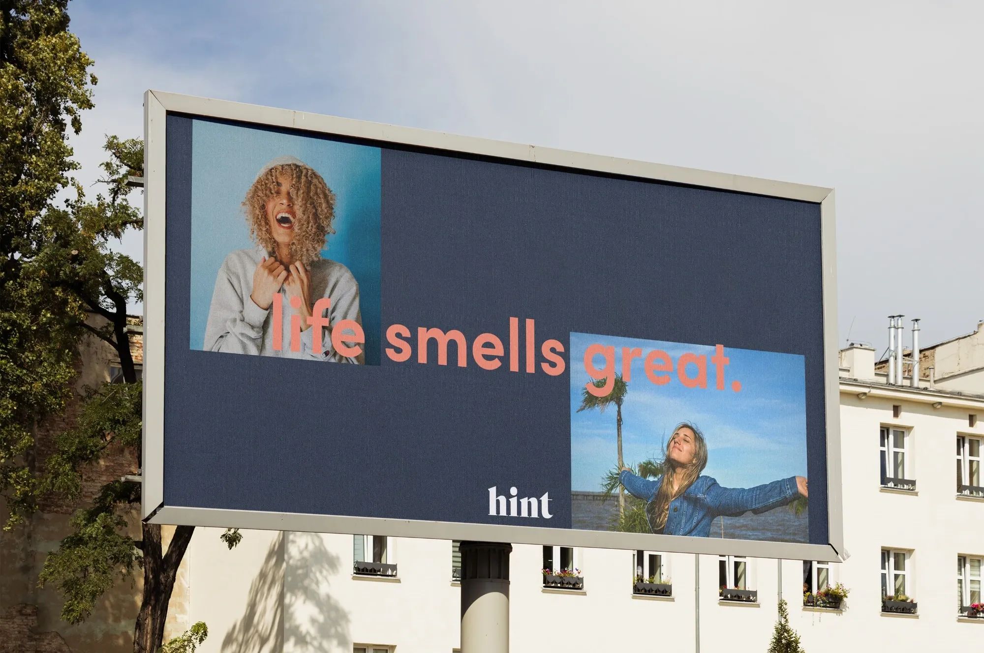 Billboard mockup for Hint with the phrase “life smells great.” in coral text, featuring joyful portraits of two women on a dark blue background.