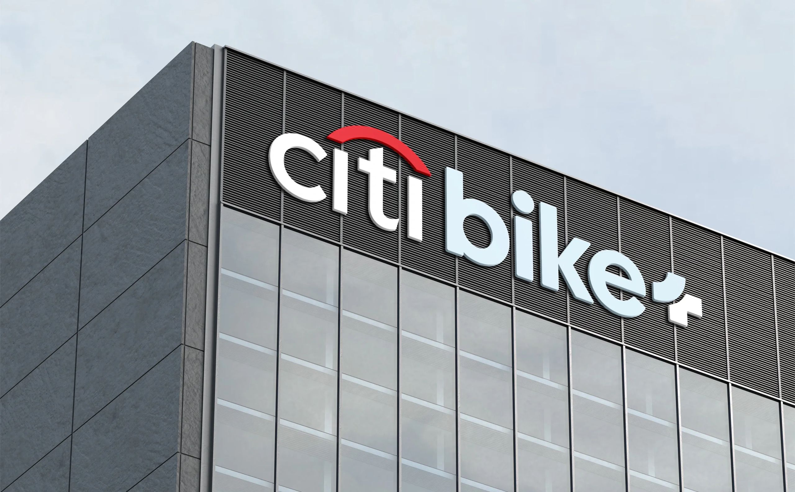 Mockup of the Citi Bike+ logo displayed on the top corner of a modern glass office building, featuring bold sans-serif lettering with the signature red arc above the "t".