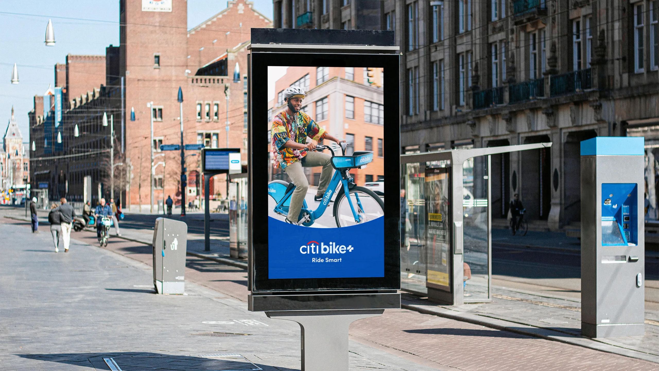 Street-level digital kiosk ad mockup for Citi Bike+ showing a smiling man riding a blue Citi Bike with the tagline “Ride Smart.” Set in an urban area with historic architecture and pedestrians in the background.