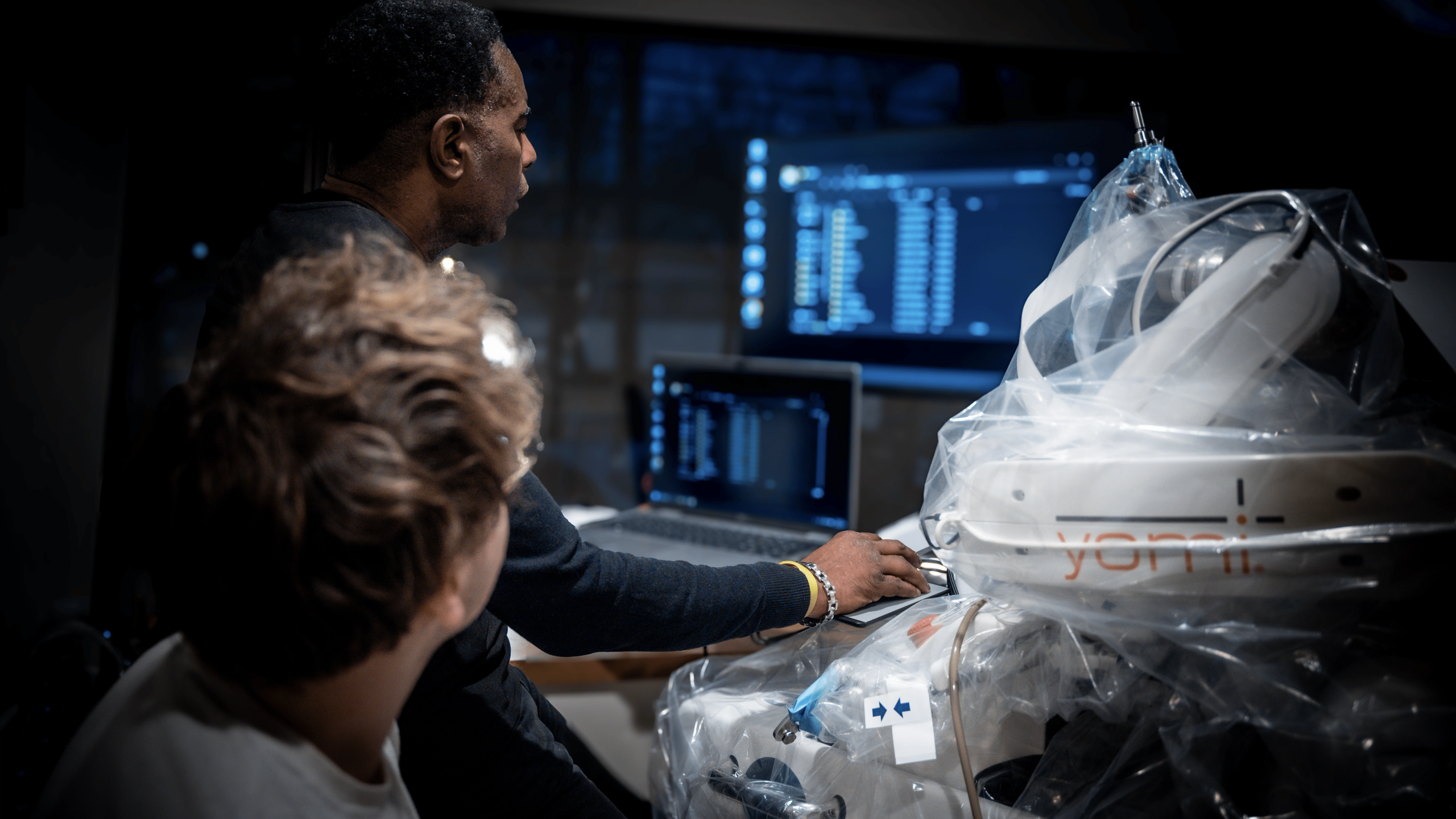 Two clinicians at a workstation beside the Yomi robotic system, with implant planning data on the monitors behind them.