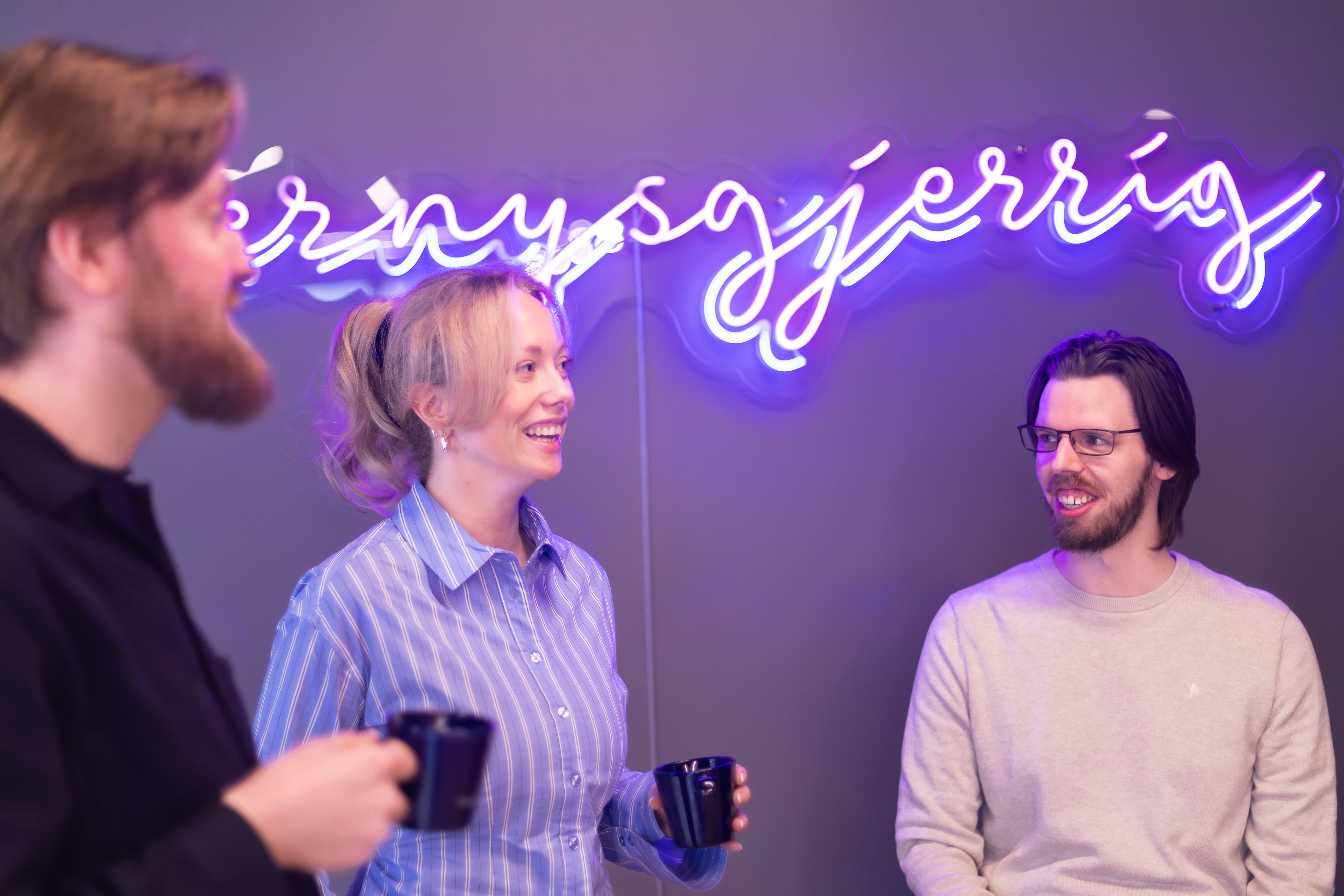 Tre kolleger med kaffekopper foran et neonskilt med ordet nysgjerrig.