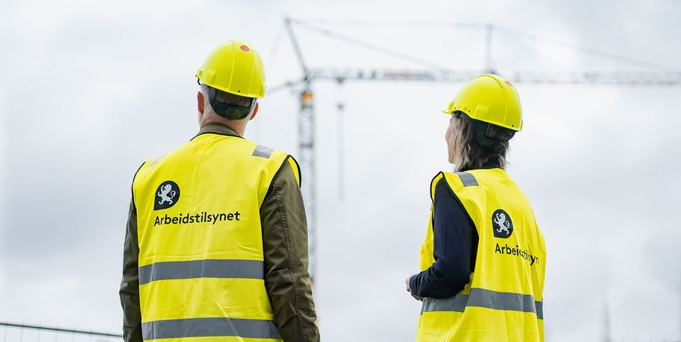 To personer fra Arbeidstilsynet med vernevest og hjelm på en byggeplass.