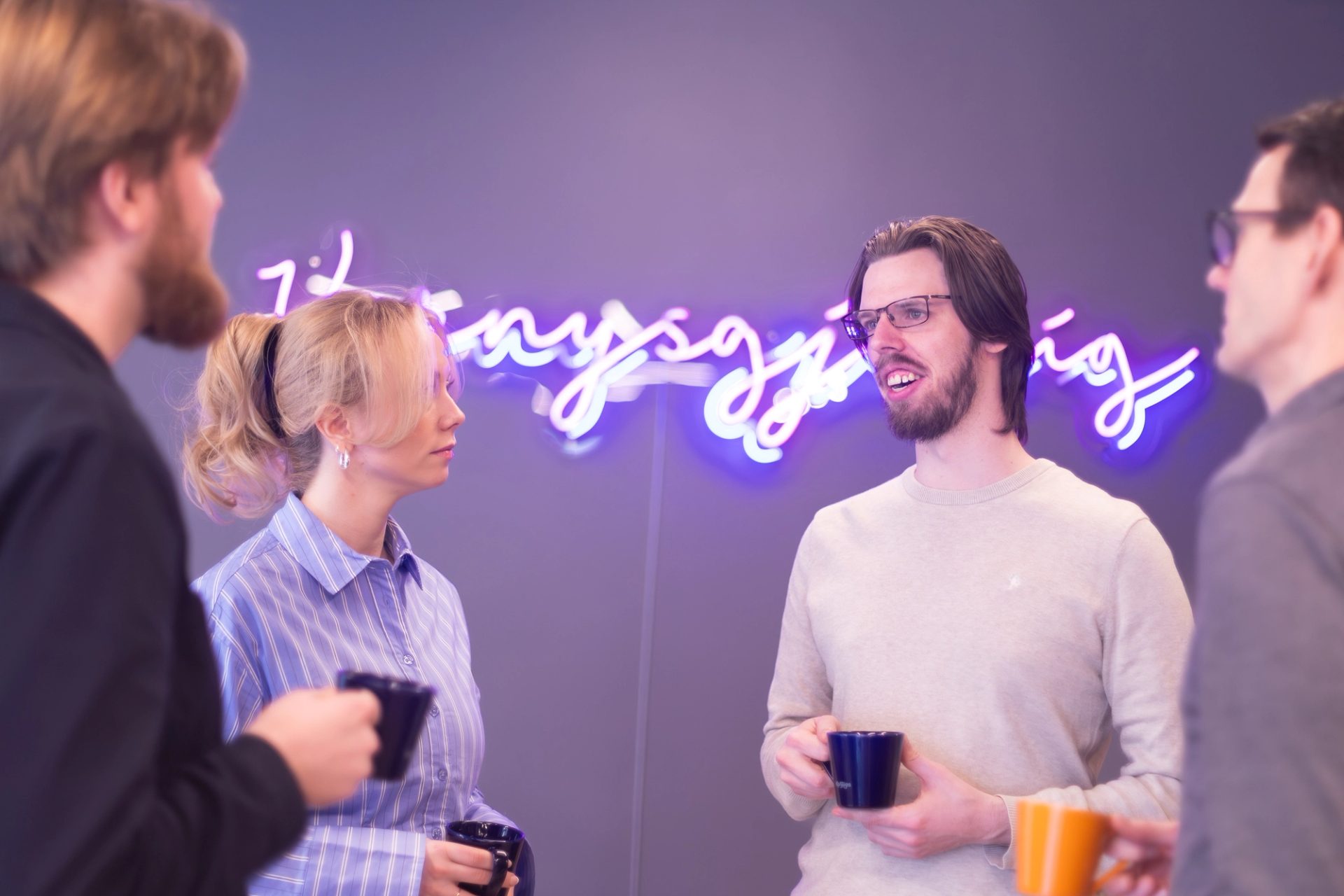 Tre Kantega-ansatte med kaffekopper under et neonskilt med ordet «nysgjerrig»