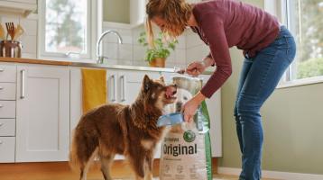 Kvinna i lila tröja matar en ivrig brun och vit hund i ett ljust kök, med en stor påse hundmat i bakgrunden.