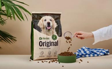 Förpackning med FirstVet Original hundfoder, en person häller foderkulor i en skål bredvid påsen. Bakgrunden inkluderar en grön växt och en blåvitrutig duk.