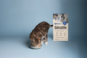 En brunfläckig katt som äter mat ur en matskål. Snett bakom står en påse kattmat av FirstVet Sensitiv. 
