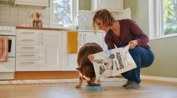 En kvinna matar sin hund i ett kök med hundfoder från en påse märkt 'FirstVet Sensitiv'. Hunden äter från en blå skål på golvet medan kvinnan häller upp maten. Köket är ljust och modernt inrett med vita skåp och stora fönster som släpper in dagsljus.