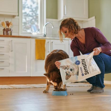 En kvinna matar sin hund i ett kök med hundfoder från en påse märkt 'FirstVet Sensitiv'. Hunden äter från en blå skål på golvet medan kvinnan häller upp maten. Köket är ljust och modernt inrett med vita skåp och stora fönster som släpper in dagsljus.