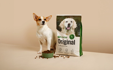 En förpackning med hundfoder från FirstVet märkt 'Original', anpassad för alla vuxna hundar. Förpackningen visar en glad golden retriever, och bredvid står en mindre hund intill en skål med torrfoder som har spillt ut några bitar. Texten på förpackningen nämner produktens egenskaper: hälsosam proteinkälla, omega-3 från fiskolja och prebiotika för en sund tarmflora.