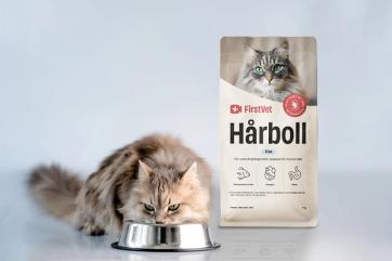 En vitaktig katt som äter mat ur en matskål. Snett bakom står en påse kattmat av FirstVet Hårboll. 