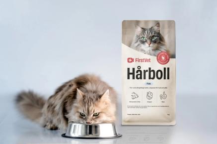 En vitaktig katt som äter mat ur en matskål. Snett bakom står en påse kattmat av FirstVet Hårboll. 