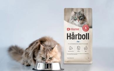 En vitaktig katt som äter mat ur en matskål. Snett bakom står en påse kattmat av FirstVet Hårboll.