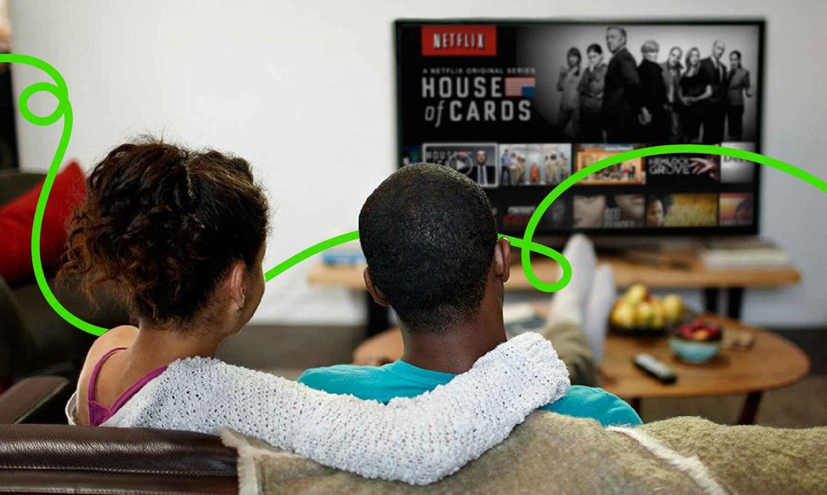 A couple watching Netflix on a couch in a cozy room; woman with her arm over man's shoulder.