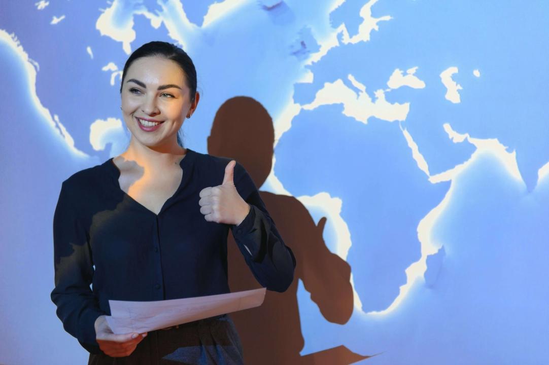 A confident woman with her hair tied back gives a thumbs-up while holding documents, standing in front of a blue-lit world map backdrop.