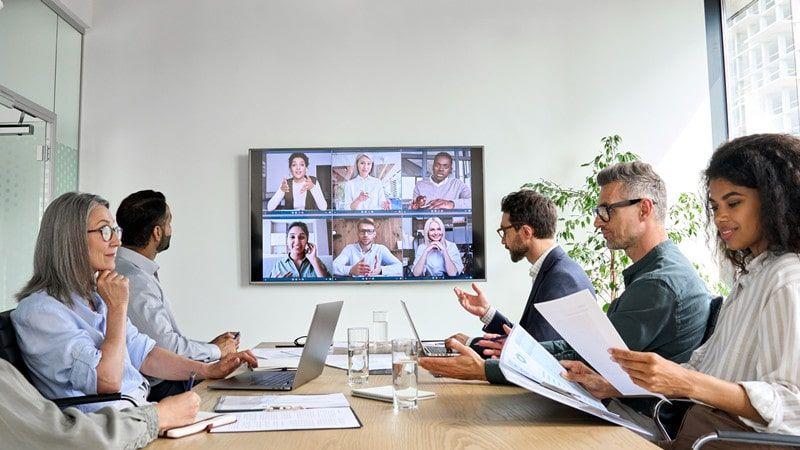 A diverse group of five professionals in a modern office, engaging in a video conference with six colleagues displayed on a large screen.