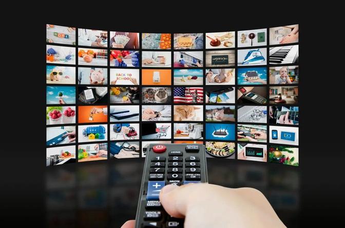 A person holding a remote control in front of a TV screen displaying multiple channels.