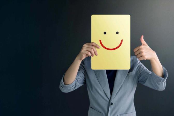 A person in a gray suit holds a yellow clipboard with a smiley face in front of their face, obscuring their identity, and gives a thumbs-up gesture with the other hand. The background is dark and plain.