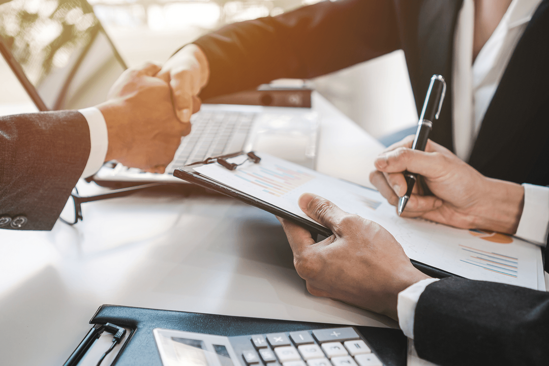Two business people shaking hands over a table with financial documents and a calculator, one person holding a pen and the other holding a clipboard.