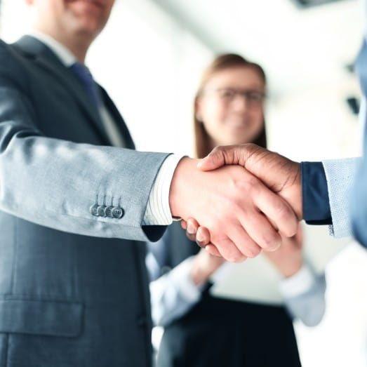 Close-up of a professional handshake between two businesspeople in a formal setting, with a smiling woman in the background, symbolizing partnership or agreement.
