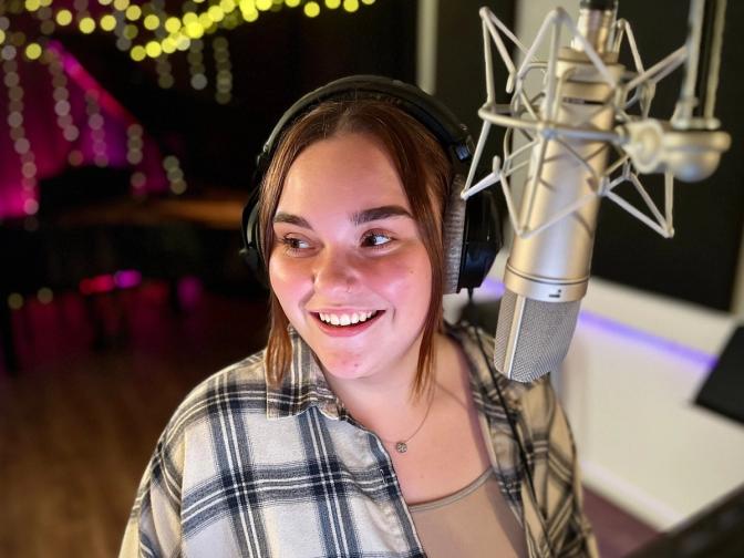 The image shows a person in a recording studio, wearing headphones and standing in front of a microphone. The studio features warm, ambient lighting and a relaxed atmosphere.