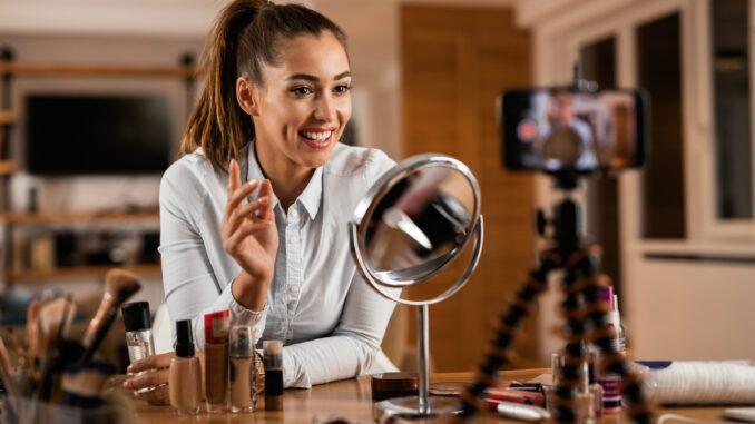 A smiling woman conducting a makeup tutorial. She's using a circular mirror while filming herself with a smartphone, surrounded by various makeup products.