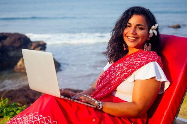 A woman in a red and white saree sits in a red chair, using a laptop by the seaside, with a flower in her hair and a cheerful smile.