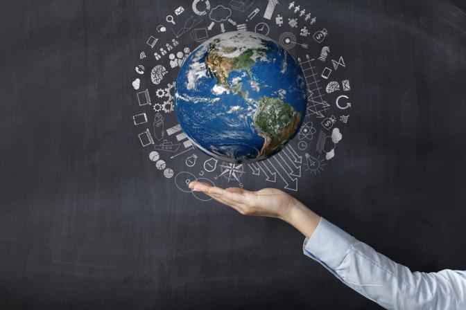 A businessperson's hand holding a globe surrounded by icons on a blackboard.