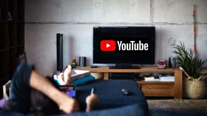 A person lying on a couch with their feet up, watching YouTube on a large TV. The YouTube logo is displayed on the screen, creating a relaxed, home-viewing atmosphere. Various items sit on a wooden table.