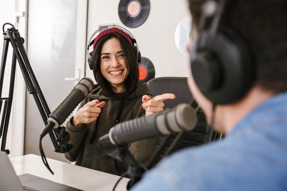 A woman wearing headphones converses with a man in a podcast recording studio.