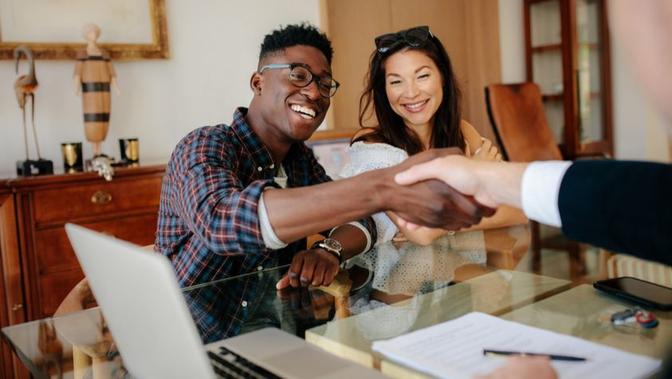 A joyful young couple shakes hands with a person across a table, sealing a deal or agreement, with laptops and documents in front of them in a bright, warmly decorated office.