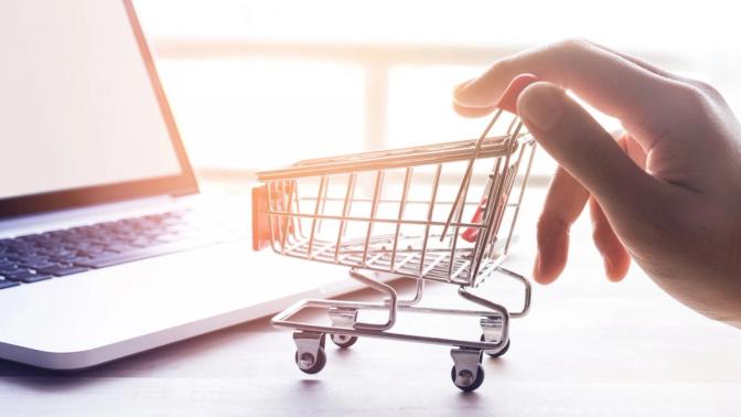 A hand pinches a miniature shopping cart, representing online shopping. In front of the hand, a computer screen suggests the digital marketplace.